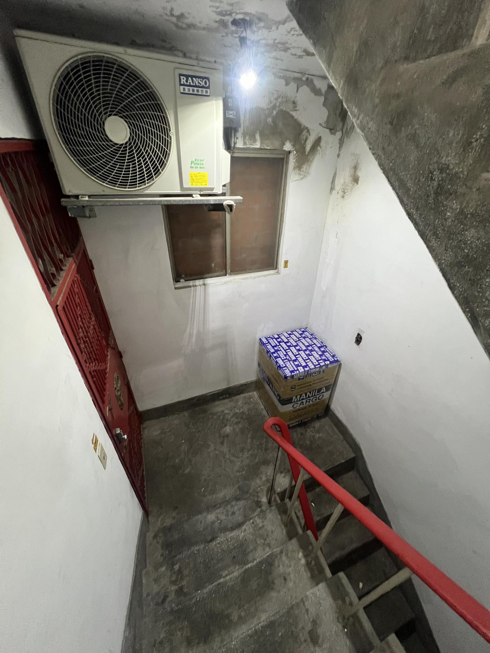 桃園某公寓的公共樓梯間牆面上方硬生生被釘上一台冷氣室外機，下方還有疑似住戶擺放的雜物箱。圖／翻攝自臉書社團「爆料公社二社」